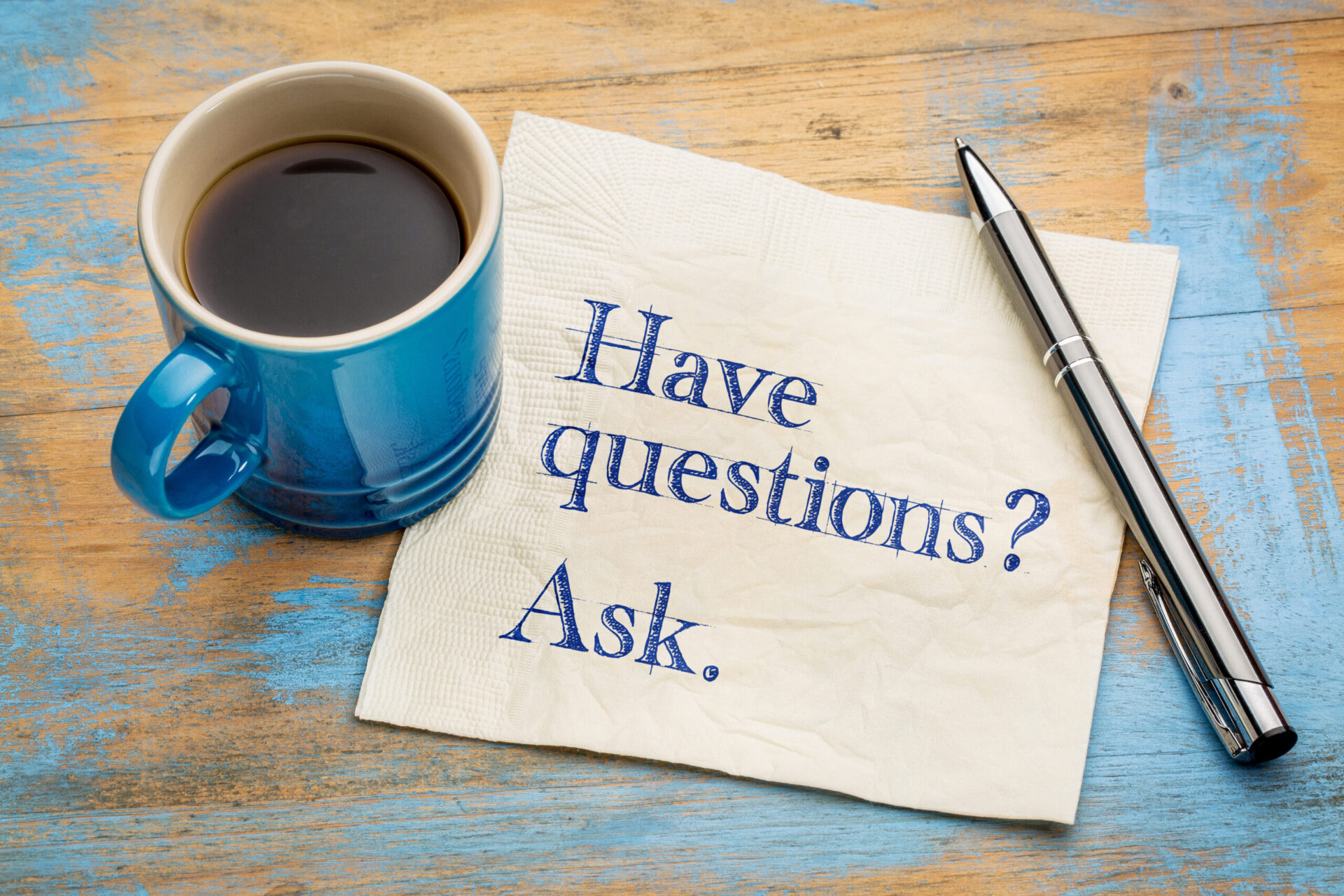 Photo of ' Have questions? Ask.' written on a napkin next to a cup of coffee